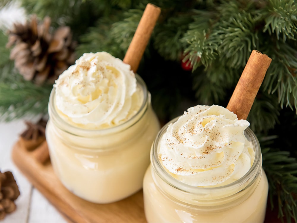 Two glasses of eggnog with whipped cream and cinnamon stick sticking out of top