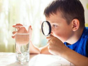 young boy inspecting glass of water on a table with a magnifying glass
