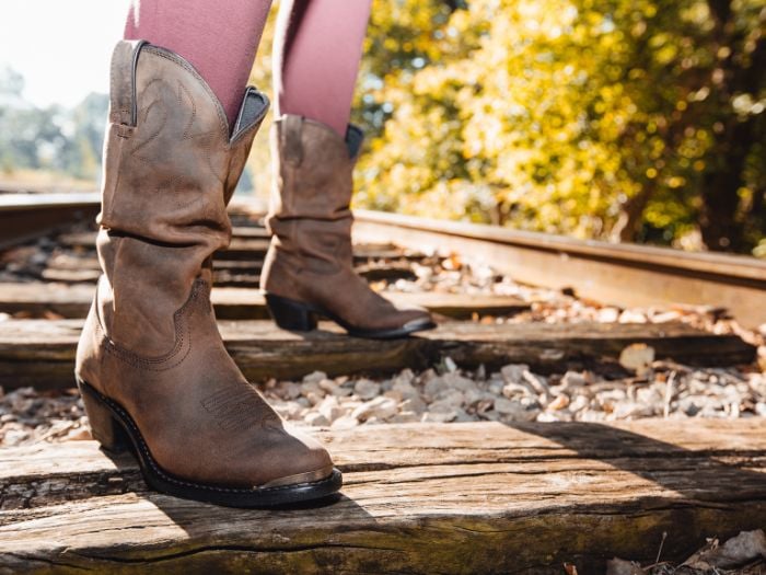 A woman wearing brown leather women western boots standing on a railroad tracks.