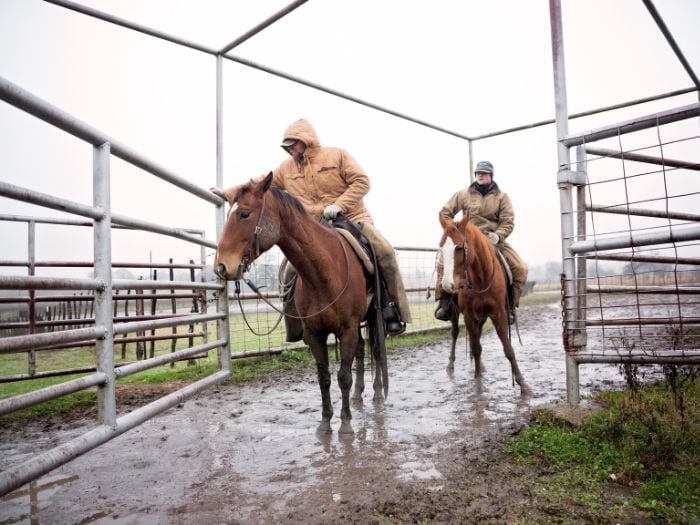 Two men riding horse through a corral in waterproof jackets.